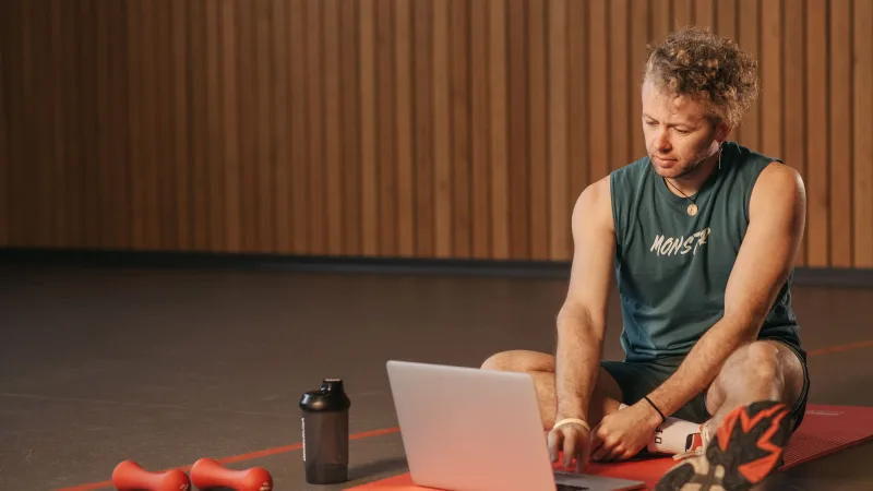 Homem sentado em tapete de exercício com laptop, cercado por halteres, cria um espaço de trabalho fitness.
